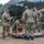 Military personnel in camouflage uniforms are conducting a combat casualty care training exercise outdoors. A simulated casualty with moulage (fake wounds/blood) is lying on a litter/stretcher while medical personnel attend to them. The scene includes a mix of U.S. Marines in MARPAT uniforms and Navy personnel in NWU Type III uniforms, with a camouflage net shelter and military tents in the background.