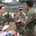 A group of military personnel in camouflage uniforms are gathered under a camouflage netting canopy in an outdoor field setting. They appear to be engaged in a briefing or training exercise, with what appears to be a medical training mannequin or casualty simulation visible in the foreground. The personnel appear to represent both U.S. Navy (NWU Type III woodland pattern) and U.S. Marine Corps (MARPAT digital pattern) uniforms.