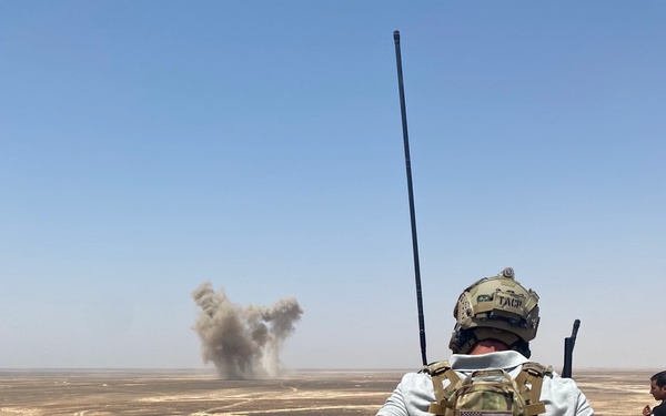 A military personnel wearing a TACP (Tactical Air Control Party) helmet and multicam plate carrier with a US flag patch observes a distant explosion or detonation in an arid desert environment. The individual is equipped with a long-whip radio antenna, suggesting they may be coordinating air or fire support. A second person is partially visible to the right.