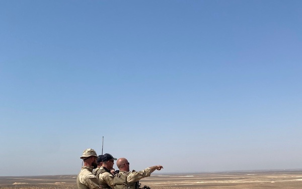 Four military personnel in camouflage uniforms are positioned on elevated rocky desert terrain, apparently conducting observation or reconnaissance activities. Three individuals are standing and one is pointing toward the distant horizon, while a fourth is kneeling behind a tripod-mounted optical/surveillance device. The landscape is an arid, flat desert extending to the horizon under a clear blue sky.