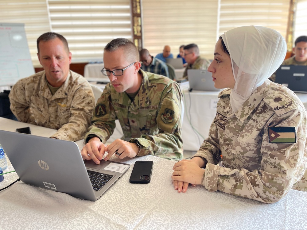 Three military personnel are seated at a table in what appears to be a joint training or briefing session. Two individuals wearing U.S. military uniforms (one in USMC desert MARPAT, one in U.S. Air Force OCP) are working on an HP laptop alongside a female soldier wearing Jordanian Armed Forces digital camouflage uniform and a white hijab. Additional military personnel are visible working in the background.