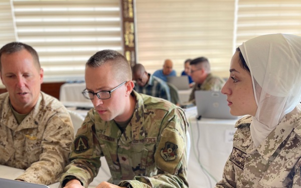Three military personnel are seated at a table in what appears to be a joint training or briefing session. Two individuals wearing U.S. military uniforms (one in USMC desert MARPAT, one in U.S. Air Force OCP) are working on an HP laptop alongside a female soldier wearing Jordanian Armed Forces digital camouflage uniform and a white hijab. Additional military personnel are visible working in the background.
