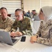 Three military personnel are seated at a table in what appears to be a joint training or briefing session. Two individuals wearing U.S. military uniforms (one in USMC desert MARPAT, one in U.S. Air Force OCP) are working on an HP laptop alongside a female soldier wearing Jordanian Armed Forces digital camouflage uniform and a white hijab. Additional military personnel are visible working in the background.