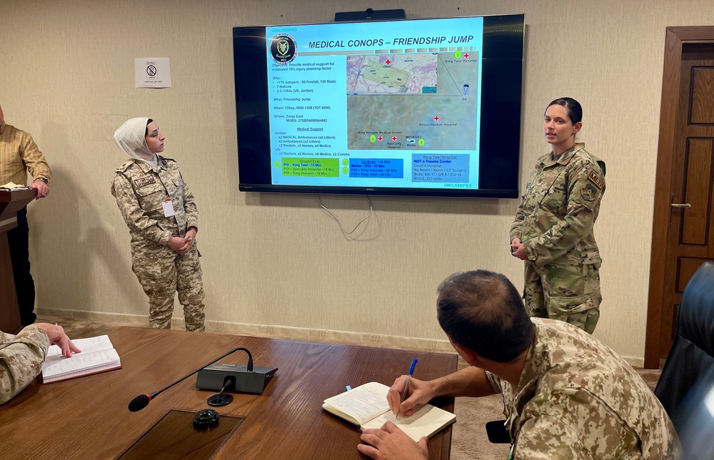 A military briefing is taking place in a conference room where personnel from what appear to be two different nations are presenting a 'Medical CONOPS – Friendship Jump' plan on a wall-mounted display screen. Two uniformed individuals stand near the screen while at least two others are seated at a conference table, with one actively taking notes. The presentation includes a tactical map, hospital locations, and medical support logistics.