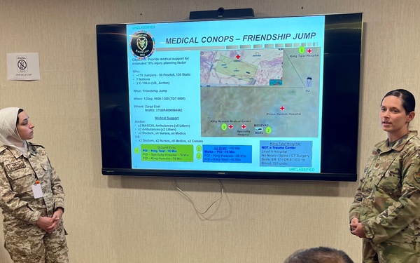 A military briefing is taking place in a conference room where personnel from what appear to be two different nations are presenting a 'Medical CONOPS – Friendship Jump' plan on a wall-mounted display screen. Two uniformed individuals stand near the screen while at least two others are seated at a conference table, with one actively taking notes. The presentation includes a tactical map, hospital locations, and medical support logistics.