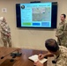 A military briefing is taking place in a conference room where personnel from what appear to be two different nations are presenting a 'Medical CONOPS – Friendship Jump' plan on a wall-mounted display screen. Two uniformed individuals stand near the screen while at least two others are seated at a conference table, with one actively taking notes. The presentation includes a tactical map, hospital locations, and medical support logistics.