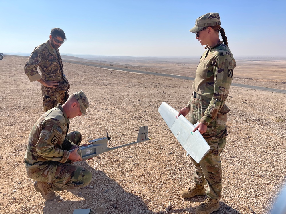 Three military personnel in camouflage uniforms are assembling or preparing a small fixed-wing unmanned aerial vehicle (UAV/drone) in an arid desert environment. Two soldiers wearing US Army OCP (Operational Camouflage Pattern) uniforms with 10th Mountain Division patches are handling the drone components, while a third individual in a different camouflage pattern observes. The setting appears to be a remote desert location with flat terrain and a road visible in the background.