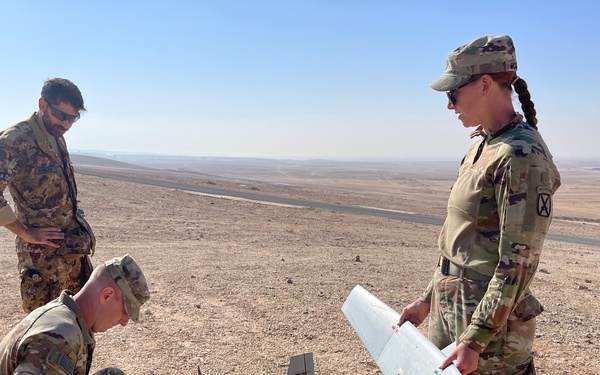 Three military personnel in camouflage uniforms are assembling or preparing a small fixed-wing unmanned aerial vehicle (UAV/drone) in an arid desert environment. Two soldiers wearing US Army OCP (Operational Camouflage Pattern) uniforms with 10th Mountain Division patches are handling the drone components, while a third individual in a different camouflage pattern observes. The setting appears to be a remote desert location with flat terrain and a road visible in the background.