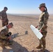 Three military personnel in camouflage uniforms are assembling or preparing a small fixed-wing unmanned aerial vehicle (UAV/drone) in an arid desert environment. Two soldiers wearing US Army OCP (Operational Camouflage Pattern) uniforms with 10th Mountain Division patches are handling the drone components, while a third individual in a different camouflage pattern observes. The setting appears to be a remote desert location with flat terrain and a road visible in the background.