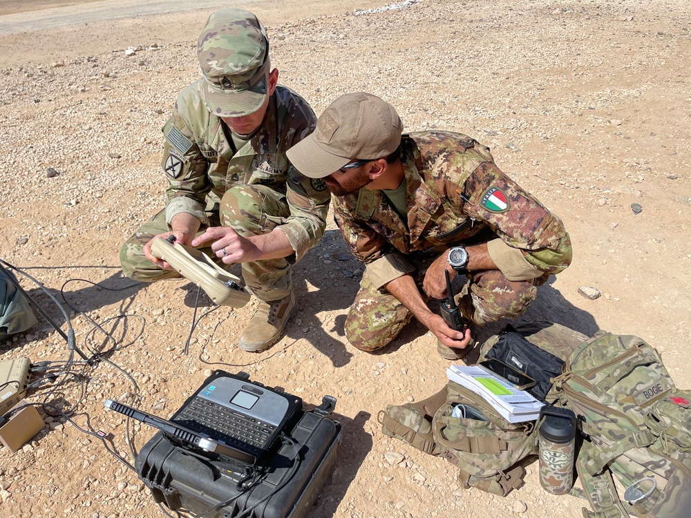 Two military personnel in camouflage uniforms are crouched on arid, rocky ground examining a piece of equipment. One soldier wears a U.S. Army uniform with a 10th Mountain Division patch and an American flag patch, while the other wears a different camouflage pattern with an Italian flag patch on the sleeve. They appear to be conducting a joint military exercise or operation involving communications or field equipment.