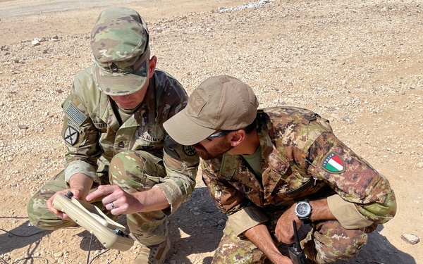 Two military personnel in camouflage uniforms are crouched on arid, rocky ground examining a piece of equipment. One soldier wears a U.S. Army uniform with a 10th Mountain Division patch and an American flag patch, while the other wears a different camouflage pattern with an Italian flag patch on the sleeve. They appear to be conducting a joint military exercise or operation involving communications or field equipment.