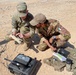 Two military personnel in camouflage uniforms are crouched on arid, rocky ground examining a piece of equipment. One soldier wears a U.S. Army uniform with a 10th Mountain Division patch and an American flag patch, while the other wears a different camouflage pattern with an Italian flag patch on the sleeve. They appear to be conducting a joint military exercise or operation involving communications or field equipment.