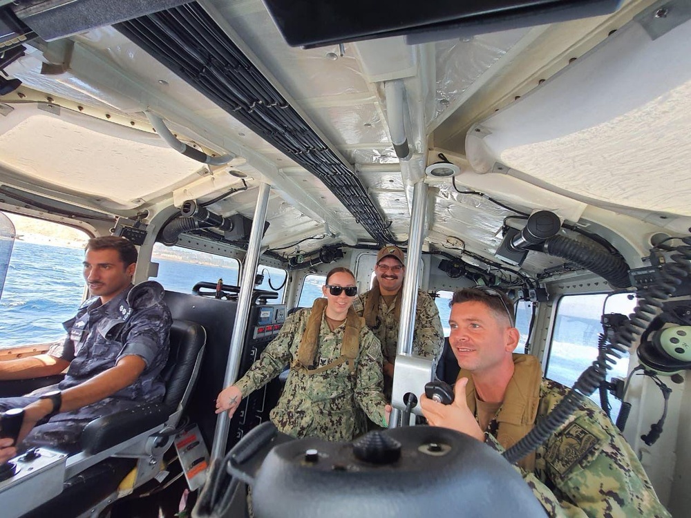 Four military personnel are seated inside the cabin of a patrol or military watercraft. Three individuals are wearing US Navy digital camouflage (NWU Type III) uniforms with tactical vests, while one person on the left wears a darker camouflage uniform suggesting a different military branch or nationality. The vessel appears to be underway, with open water and a coastline visible through the windows.