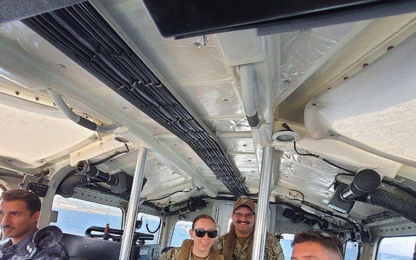 Four military personnel are seated inside the cabin of a patrol or military watercraft. Three individuals are wearing US Navy digital camouflage (NWU Type III) uniforms with tactical vests, while one person on the left wears a darker camouflage uniform suggesting a different military branch or nationality. The vessel appears to be underway, with open water and a coastline visible through the windows.