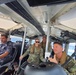 Four military personnel are seated inside the cabin of a patrol or military watercraft. Three individuals are wearing US Navy digital camouflage (NWU Type III) uniforms with tactical vests, while one person on the left wears a darker camouflage uniform suggesting a different military branch or nationality. The vessel appears to be underway, with open water and a coastline visible through the windows.