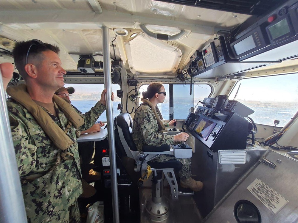Three military personnel in U.S. Navy digital camouflage uniforms are aboard the bridge/helm station of a patrol or coastal vessel underway on open water. One sailor is seated at the helm operating navigation controls, while two others stand and observe. The vessel's interior is equipped with multiple electronic navigation and communication systems.