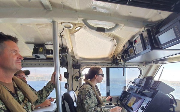 Three military personnel in U.S. Navy digital camouflage uniforms are aboard the bridge/helm station of a patrol or coastal vessel underway on open water. One sailor is seated at the helm operating navigation controls, while two others stand and observe. The vessel's interior is equipped with multiple electronic navigation and communication systems.