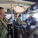 Three military personnel in U.S. Navy digital camouflage uniforms are aboard the bridge/helm station of a patrol or coastal vessel underway on open water. One sailor is seated at the helm operating navigation controls, while two others stand and observe. The vessel's interior is equipped with multiple electronic navigation and communication systems.