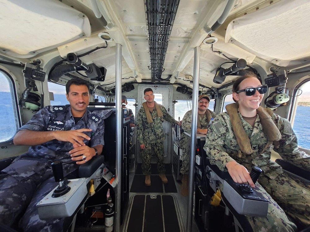 The image shows military personnel from at least two different nations aboard what appears to be a naval patrol vessel or small military watercraft. Personnel in U.S. Navy digital camouflage (NWU Type III) are seated at helm controls alongside an individual in a darker naval uniform with Arabic script insignia, suggesting a joint or cooperative naval exercise. The vessel interior shows navigation and control equipment, and water is visible through the windows.