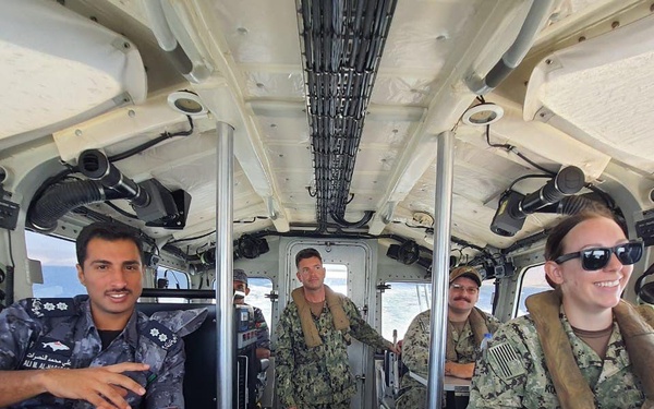 The image shows military personnel from at least two different nations aboard what appears to be a naval patrol vessel or small military watercraft. Personnel in U.S. Navy digital camouflage (NWU Type III) are seated at helm controls alongside an individual in a darker naval uniform with Arabic script insignia, suggesting a joint or cooperative naval exercise. The vessel interior shows navigation and control equipment, and water is visible through the windows.