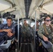 The image shows military personnel from at least two different nations aboard what appears to be a naval patrol vessel or small military watercraft. Personnel in U.S. Navy digital camouflage (NWU Type III) are seated at helm controls alongside an individual in a darker naval uniform with Arabic script insignia, suggesting a joint or cooperative naval exercise. The vessel interior shows navigation and control equipment, and water is visible through the windows.