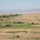 Three military attack helicopters, identifiable as AH-64 Apache variants, are flying in a low-altitude formation over a semi-arid, hilly landscape. The terrain features dry grassland, scattered trees, and small rural structures in the background. Mountains are visible on the horizon under a hazy sky.
