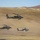 Three military attack helicopters are flying in close formation at low altitude over an arid, desert-like terrain. The helicopters appear to be AH-64 Apache attack helicopters, identifiable by their distinctive tandem-seat cockpit and stub-wing weapons pylons. Rotor wash is visibly kicking up dust from the dry ground below, suggesting the aircraft are operating at very low altitude.