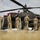 A group of U.S. Army soldiers in MultiCam/OCP camouflage uniforms are gathered around an AH-64 Apache attack helicopter named 'Miss Mabel' on a gravel/dirt landing area. The personnel appear to be conducting pre- or post-flight maintenance, inspection, or a briefing around the aircraft. The scene takes place in an open field environment under overcast skies.