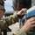 A military service member in MultiCam/OCP camouflage uniform is working on what appears to be an aircraft weapons or ammunition loading system. The individual is wearing hearing protection (ear defenders) and glasses while carefully handling rounds or cartridges arranged in a linked belt or magazine assembly. The setting appears to be an outdoor flight line or airfield.