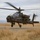 A military attack helicopter, identifiable as an AH-64 Apache, is captured in a low hover or during takeoff/landing over a dry, grassy field. The aircraft is fully armed with visible weapon systems and displays unit insignia on its fuselage. The rotor blades show significant motion blur, indicating active flight operations.