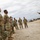 A group of U.S. Army soldiers in Operational Camouflage Pattern (OCP) uniforms are gathered outdoors in what appears to be an arid, open area. One soldier on the right is addressing the group with a hand gesture, suggesting a briefing or after-action review. Military helicopters, likely Apache attack helicopters, are visible in the background.