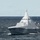 A modern stealth corvette with angular, low-observable hull design is underway on open ocean waters. The vessel displays a distinctive geometric camouflage pattern in shades of gray and features a pyramidal mast structure characteristic of stealth warship design. A hull number beginning with 'K35' is partially visible on the bow.