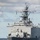 A U.S. Navy warship bearing hull number '44' is underway at sea under partly cloudy skies. The vessel is a large amphibious or surface combatant ship viewed from a bow-on perspective, showing extensive superstructure, radar arrays, and communications equipment. An American flag and what appears to be a signal flag are visible on the mast.
