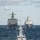 Three naval warships are sailing in formation on open ocean under a partly cloudy sky. The foreground vessel is a smaller grey warship flying what appears to be a Swedish flag, while two larger US Navy vessels — identifiable by their grey hulls and distinct superstructures — follow in the background. The scene suggests a multinational naval exercise or transit operation.
