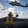 A U.S. Navy Landing Signal Enlisted (LSE) crew member aboard what appears to be USS Kearsarge (LHD-3) guides a helicopter during a vertical replenishment (VERTREP) operation at sea. An MH-60S Seahawk helicopter hovers overhead, lowering a cargo net containing supplies to the flight deck. The operation is taking place in open ocean under partly cloudy skies.