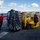 A yellow naval forklift is moving a large netted cargo pallet on the flight deck of a U.S. Navy ship at sea. Two personnel in red jackets and protective gear are managing the cargo transfer operation. The scene appears to be a replenishment-at-sea (RAS) or vertical replenishment (VERTREP) evolution.