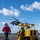 A military helicopter, appearing to be an MH-60 Seahawk, is hovering or landing on the flight deck of a U.S. Navy vessel. A deck crew member in a red jersey and helmet is walking toward the aircraft, while another crew member in green operates a yellow Liftking forklift/deck equipment vehicle marked 'USYN-3503'. The scene depicts active flight deck operations at sea under partly cloudy skies.