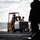 A yellow U.S. Navy forklift marked 'USN 13-64197' is transporting a shrink-wrapped pallet of supplies across what appears to be the flight deck of a naval vessel. At least two personnel wearing helmets and colored jerseys are visible — one operating the forklift and one standing nearby. The ocean is visible in the background, confirming an at-sea replenishment or cargo-handling operation.
