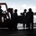 The image shows a group of personnel working on what appears to be a naval vessel's flight deck or hangar bay, handling cargo boxes with the assistance of a forklift. The workers are wearing helmets and colored jerseys/vests, consistent with U.S. Navy flight deck crew attire. The ocean is visible in the background, confirming a maritime setting.