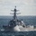 A U.S. Navy Arleigh Burke-class guided-missile destroyer bearing hull number 117 is underway at sea in moderately rough conditions. The vessel is cutting through choppy waves, generating significant bow spray, and appears to be traveling at a notable speed. The overcast sky and whitecapped seas suggest active weather conditions during this transit.