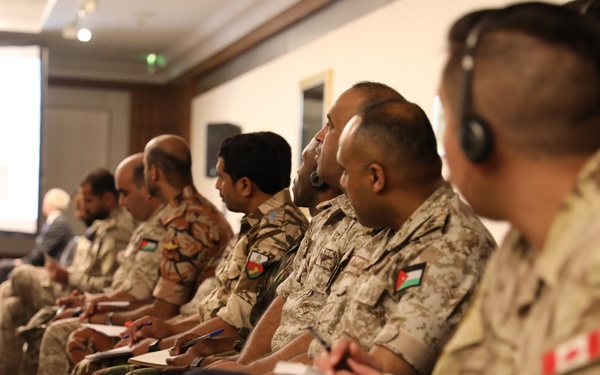 A group of military personnel from multiple nations are seated in rows attending a formal briefing or conference in what appears to be a hotel or conference room setting. Participants are wearing various camouflage uniforms with visible national flag patches, including Jordanian, Canadian, and other Middle Eastern nation insignia. Several attendees are taking notes while facing a projected presentation screen at the front of the room.
