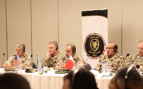 A multinational military conference is taking place at the 'Eager Lion 2022' exercise, as indicated by the banner in the background. Military officers from multiple nations are seated at a long conference table wearing translation headsets, with small national flags visible including the United States, Bahrain, Saudi Arabia, and Jordan. The foreground shows additional uniformed personnel also wearing headsets, suggesting a large multilateral military coordination meeting.