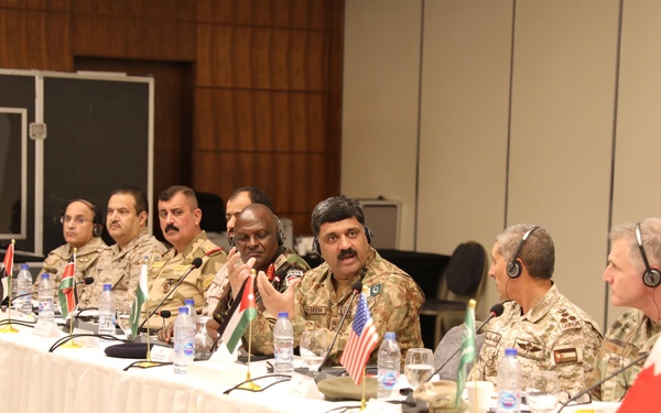 A multinational military conference or meeting is taking place at a long conference table draped in white cloth. Military officers in various uniforms are seated together, with small national flags identifying their respective countries placed on the table. The setting appears to be a formal diplomatic or strategic military coordination meeting.