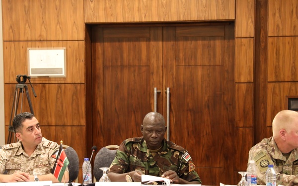 Three military officers from different nations are seated at a conference table during what appears to be a multinational military meeting or summit. The officers represent Jordan, Kenya, and the United States, as indicated by name placards and national flags visible on the table. The setting is a formal conference room with wood-paneled walls.