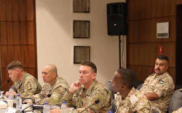 A multinational military conference or briefing is taking place at a formal meeting table. Personnel from at least three nations are present, identifiable by visible flag patches including the United States, Canada, and Jordan. The setting appears to be a conference room with microphones, name placards, and water bottles arranged along the table.