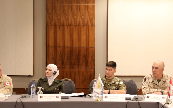 Four military personnel from different nations are seated at a conference table during what appears to be a multinational military meeting or briefing. Name placards and small national flags are visible on the table, indicating a formal international military conference setting. The participants are wearing different national military uniforms and appear to be listening or attending a presentation.