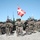 A group of military personnel in full combat gear pose for a formation photograph in an arid, rocky terrain. Two flags are visible — a Danish flag (Dannebrog) prominently in the center and a United States flag to the left — suggesting a joint multinational military exercise or operation. Several armored military vehicles, including what appear to be Stryker-type wheeled armored personnel carriers, are positioned in the background.