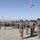 A multinational military formation ceremony is taking place on a gravel parade ground under clear blue skies. Multiple national flags are visible, including what appears to be the Georgian flag (white with red crosses), the US flag, and other national banners. Soldiers in various camouflage uniforms are standing in formation, with some carrying flags and weapons.