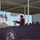 A woman in a red jacket is delivering a speech at a podium during the 'Noble Partner' military exercise event. The podium displays the Noble Partner logo featuring combined US and Georgian emblems. Multiple national flags are visible in the background alongside military personnel.