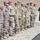 A military formation ceremony is taking place outdoors on a gravel surface, featuring soldiers from multiple nations standing at attention in a line. The soldiers wear various camouflage uniforms with different national patches and beret colors, indicating a multinational military event. A civilian in traditional dark attire stands to the right, holding what appears to be a folded flag or ceremonial item.
